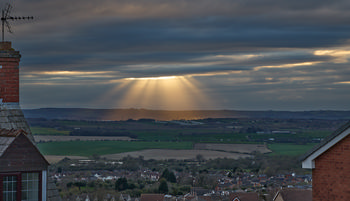 This landscape photograph captures a rural scene in Derbyshire, United Kingdom, during a spring evening as witnessed from the hillside houses of Bolsover in England. The main feature of the image is the striking display of God's rays, also known as crepuscular rays, streaming dramatically through breaks in the clouds and illuminating the distant landscape. Below this natural phenomenon, a solar farm with clearly visible rows of solar panels stretches across the fields, highlighting the blend of natural beauty and renewable energy within the region. The houses in the foreground emphasize the rural community setting, while the rolling hills and patchwork fields of Derbyshire serve as a picturesque backdrop enhanced by the soft, diffused light of the spring evening.